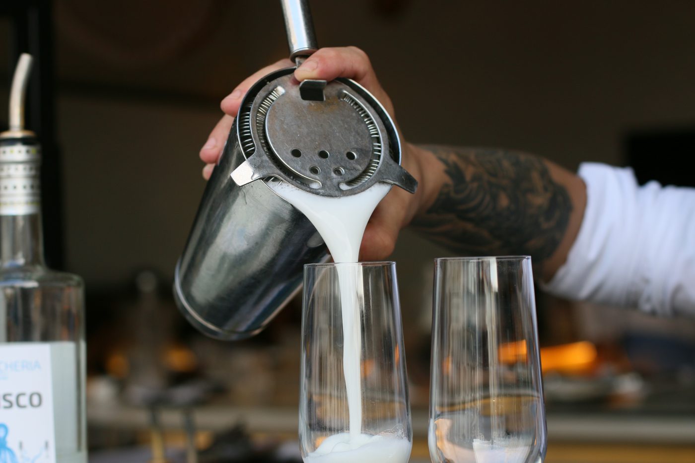 Barkeeper gießt einen frisch geschüttelten Pisco Sour aus dem Shaker in zwei Gläser, daneben eine Flasche Pisco